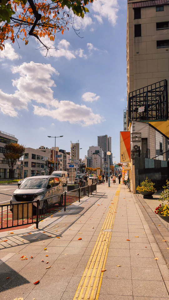 Sunny autumn day on a bustling Japanese city street lined with buildings and cars, under a bright blue sky.